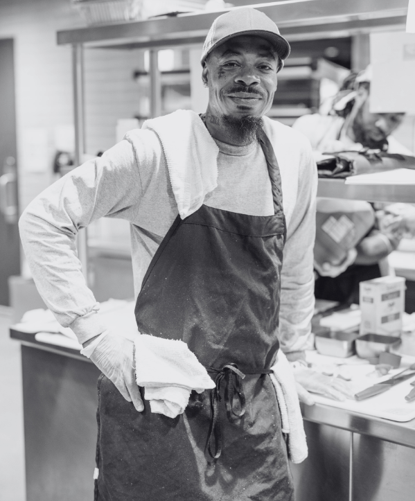 African American man standing in a kitchen and grinning. He is wearing an apron and a hat.